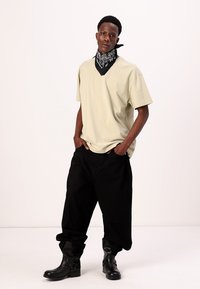 T-shirt oversized beige, pantalon baggy noir, et bottes noires élégantes. Un bandana noir autour du cou arbore un motif paisley blanc.