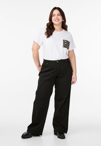 Valkoinen t-paita, jossa on leopardikuosiset taskut, yhdistettynä korkeavyötäröisiin mustiin leveälahkeisiin housuihin ja massiivisiin mustiin kenkiin. Yksinkertainen, rento asu.