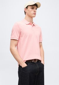 Νεαρός άνδρας που φοράει ανοιχτό ροζ πόλο πουκάμισο, σκούρο τζιν παντελόνι, μπεζ καπέλο και καφέ ζωνή, με τα χέρια στις τσέπες, κοιτάζοντας προς τα εμπρός.