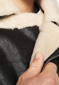 Czarna skórzana kurtka o teksturowej powierzchni, z kremowym futrzanym podszyciem i widocznym zamkiem. Ręka trzymająca brzeg kurtki.