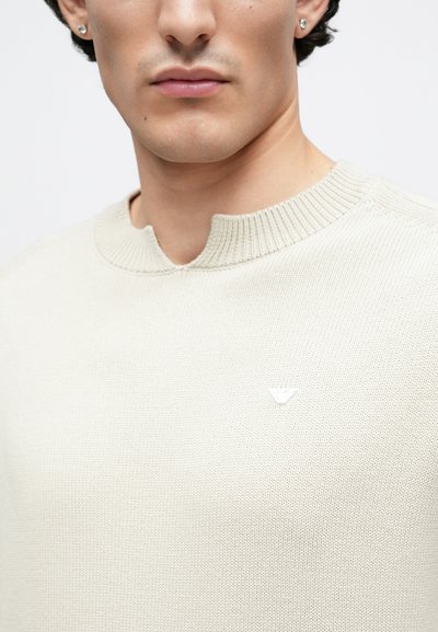 Μπεζ πλεκτό πουλόβερ με στρογγυλή, ριμπ λαιμόκοψη και μικρό λευκό λογότυπο στο στήθος. Υφασμάτινη υφή με λείο φινίρισμα.