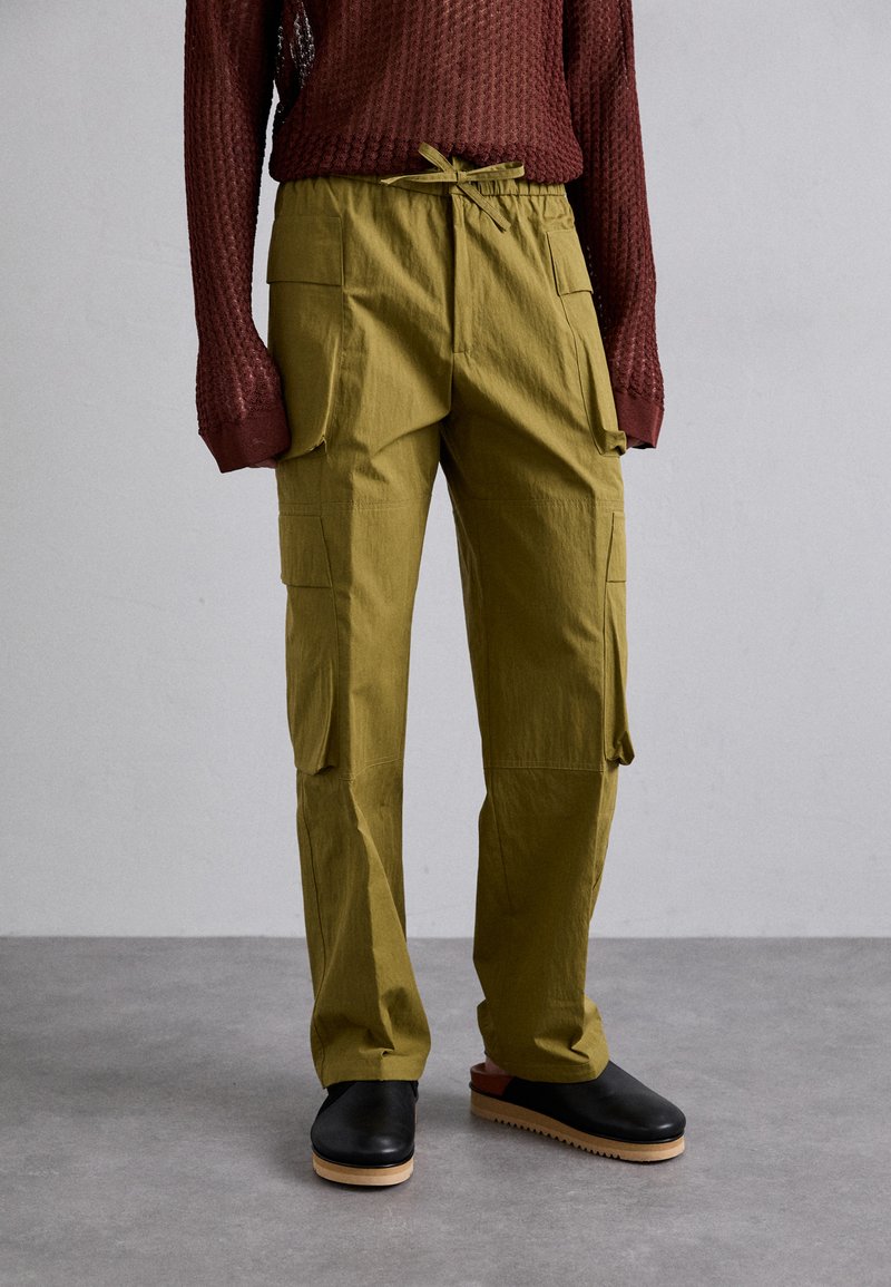 Olive green cargo pants with side pockets, elastic waistband, and drawstring. Paired with black clogs featuring a light wooden sole.
