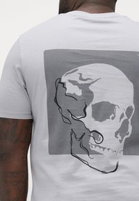 Lys grå bomull t-skjorte med et svart-hvitt hodeskallegrafikk på baksiden, satt mot en mørkere grå rektangulær bakgrunn.