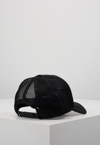 Casquette trucker noire en mesh avec fermeture snapback, visière légèrement incurvée, posée sur une surface blanche sur fond gris.