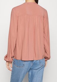 Blouse à manches longues, couleur rose poussiéreux, avec un détail froncé au dos et un tissu doux. À associer avec un jean bleu clair, mettant en valeur des manches élastiques.