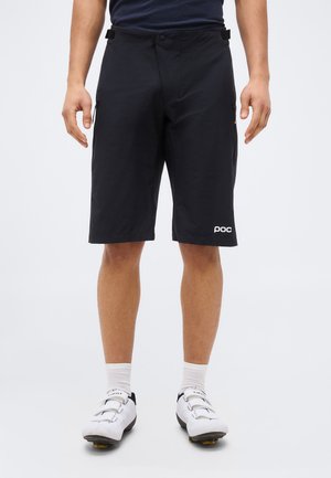 Man draagt zwarte fietsbroeken, witte sokken en witte fietsschoenen, staand tegen een effen lichte achtergrond.