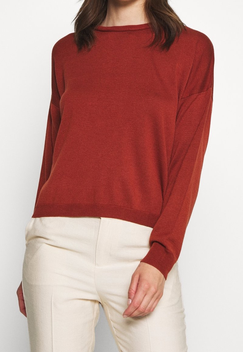 Femme portant un pull ample à manches longues couleur rouille et un pantalon crème taille haute sur fond blanc.