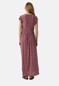 Robe maxi de couleur violet foncé avec un motif floral, manches flottantes courtes, taille smockée et tissu fluide. Portée avec des sandales décontractées.