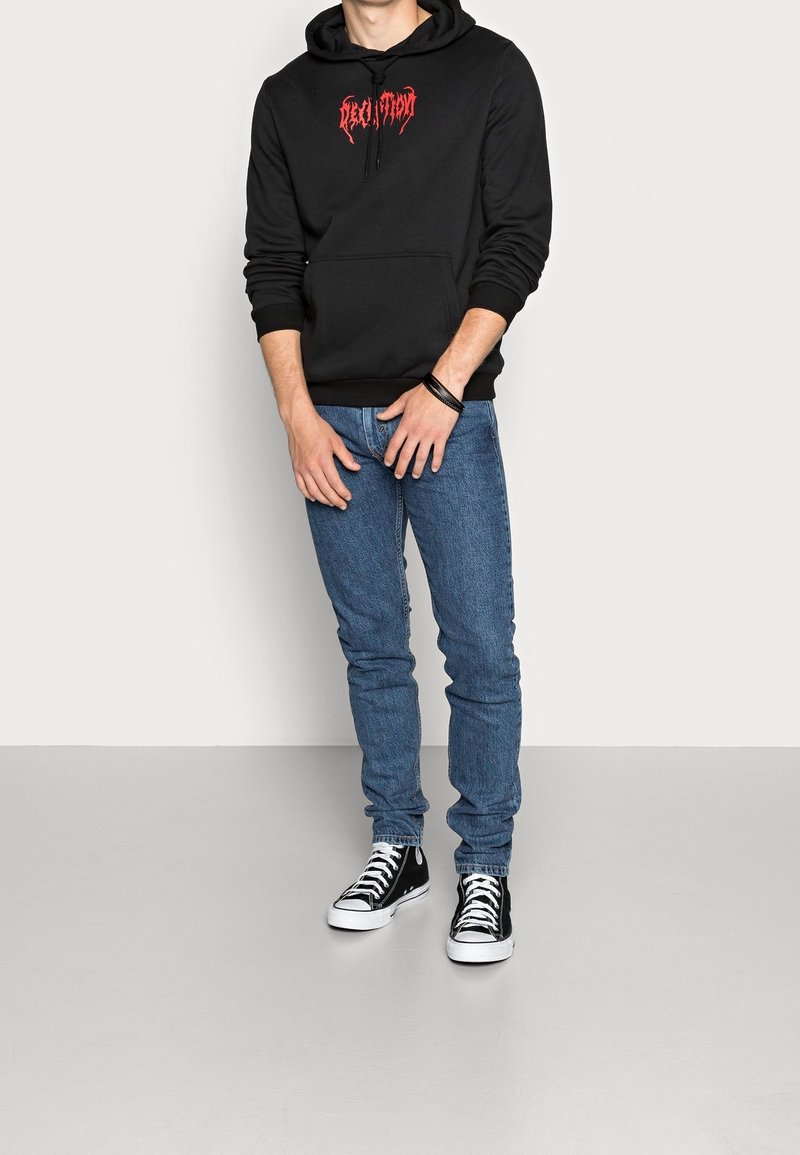 Jeune personne portant un sweat à capuche noir avec un texte rouge, un jean bleu, des baskets noir et blanc, et un bracelet noir, debout devant un fond uni.