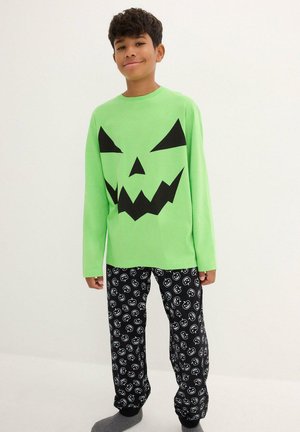 La camicia a maniche lunghe di colore verde presenta un grande volto di zucca nera; abbinata a pantaloni neri con illustrazioni di zucche bianche. Tessuto morbido, vestibilità comoda.