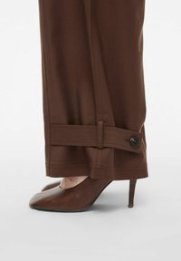Hnědé tailored kalhoty s detailem na manžetách na knoflíky, zkombinované s hnědými koženými jehlovými podpatky, viditelné z boku. Hladká textura, elegantní design.