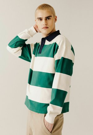 Jeune personne aux cheveux blonds courts portant une chemise à manches longues rayée verte et blanche avec un col noir, posant avec une main derrière la tête.