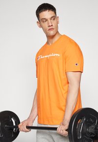 Orange Baumwoll-T-Shirt mit einem weißen "Champion"-Logo, kurzen Ärmeln und einem runden Halsausschnitt, getragen von einer Person, die eine schwarze Hantel hält.