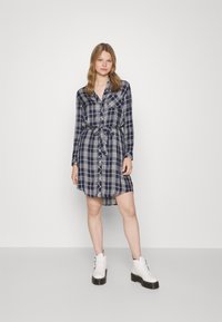 Abito camicia a quadri navy con maniche lunghe, colletto, chiusura a bottoni sul davanti e cintura in vita. Indossato con stivali bianchi alla caviglia con fibbie.