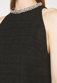 Gros plan sur une robe noire texturée sans manches, avec un col montant orné de strass argentés.
