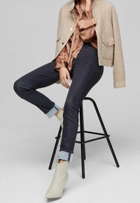 Chaqueta corta de tweed beige, blusa de satén marrón, jeans oscuros con puños azul claro y botas de tobillo crema con punta afilada, sentada en un taburete negro.