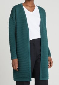 Teal ribbad kofta med långa ärmar, öppen front och texturerad tyg. Bärs över en vit V-ringad skjorta och matchas med mörka byxor.