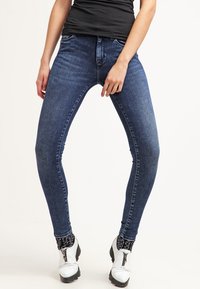 Jeans ajustados de mezclilla azul oscuro, con un diseño clásico de cinco bolsillos, ajuste ceñido y un ligero desgaste en la parte frontal.