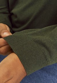 Ciemnozielony dziergany sweter z detalem prążkowanych mankietów, ukazujący miękką fakturę. Widoczna ręka trzymająca mankiet, na tle niebieskiego denimu.