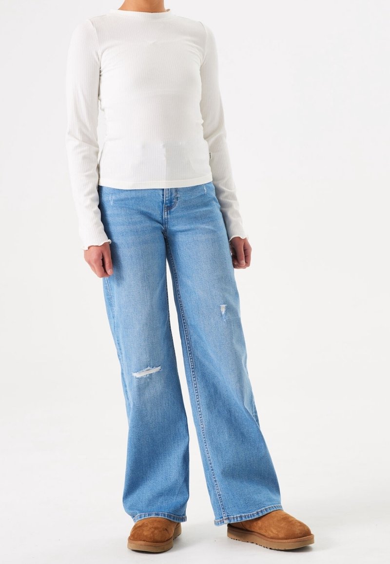 Personne portant un haut blanc à manches longues, un jean bleu clair déchiré à jambes larges, et des chaussures marron en daim sans lacets, debout devant un fond blanc.