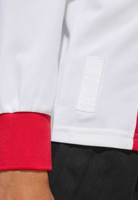 Biela športová košeľa s červenými manžetami, vyrobená z textúrovanej sieťoviny a s obdĺžnikovou embosovanou nálepkou na boku.