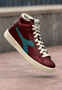 High-top sneaker in burgundy leather with teal accents, white laces, and a textured cream rubber sole. Features logo on the tongue.