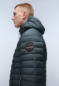 Giacca imbottita blu scuro con cappuccio, texture trapuntata, zip laterali e un patch rotondo con logo sulla spalla sinistra.