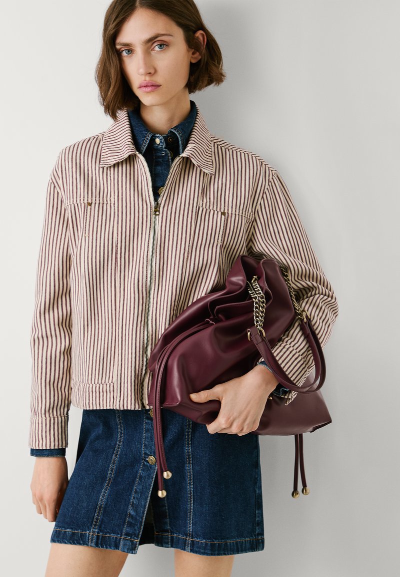 Casaco de ganga às riscas com linhas vermelhas claras e escuras, combinado com uma saia de ganga escura. Segurando uma mala de couro bordô com detalhes em corrente.