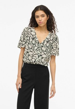 Femme aux cheveux bouclés et foncés portant un chemisier beige avec des contours floraux noirs et un pantalon noir taille haute, debout devant un fond blanc.