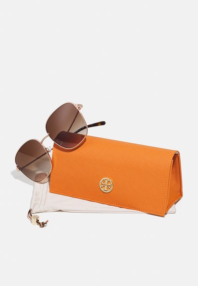 Occhiali da sole color marrone con montatura in metallo dorato, insieme a un astuccio arancione brillante e texture con logo dorato. Entrambi gli oggetti sono su uno sfondo bianco.