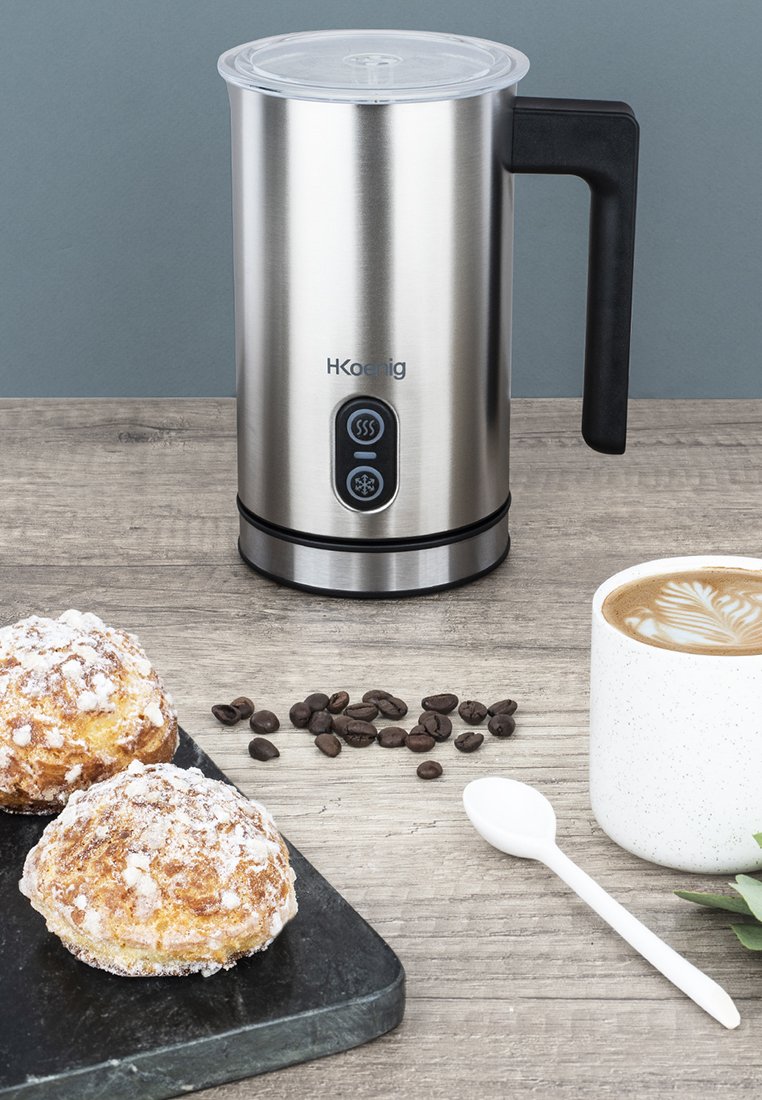 Frullatore per latte in acciaio inossidabile con manico nero, due pulsanti per caldo e freddo, accompagnato da una tazza di caffè, pasticcini e chicchi di caffè.