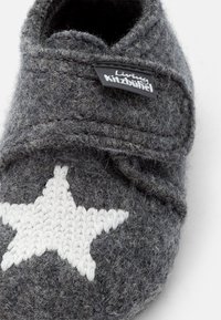 Gråfiltad toffel med ett stickat vitt stjärndetalj, med en strukturerad yta och en svart etikett på sidan. Enkelt design för inomhusbruk.