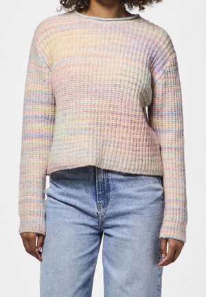 Femme portant un pull tricoté à rayures multicolores pastel à manches longues et un jean bleu clair taille haute.