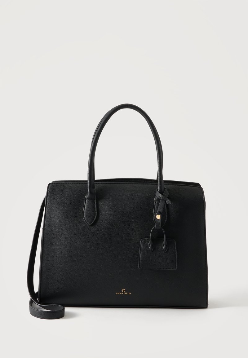 Sac à main en cuir structuré noir avec deux poignées, une bandoulière amovible et une petite étiquette attachée sur fond blanc.