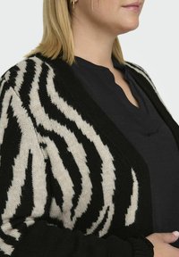 Zwart en crèmekleurig gebreid cardigan met zebra-strepen, met een gestructureerde afwerking, open voorkant en lange mouwen, gedragen over een donkere top.