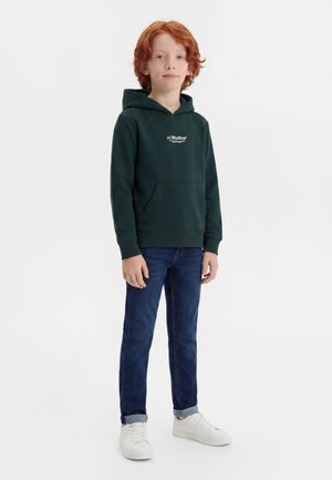 Garçon roux debout, portant un sweat à capuche vert foncé avec une poche avant, un jean bleu retroussé aux chevilles et des baskets blanches.
