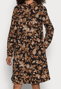 Svart klänning med ett blommönster i orange, kräm och brunt, med lång ärm, krage, knäppning med knappar och en avslappnad passform.