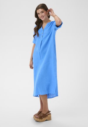 Mosolygó nő kék midi ruhában, 3/4-es ujjú, barna platform szandálban, egyszerű világos háttér előtt.