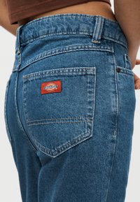 Jean en denim bleu taille haute, avec un patch logo Dickies rouge sur la poche arrière et des détails de couture visibles.