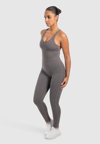 Kvinde står oprejst, iført en tætsiddende grå ærmeløs jumpsuit og hvide sneakers, med håret sat op i en knold, foran en ensfarvet lys baggrund.