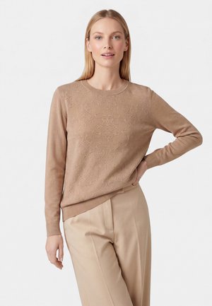 Donna con capelli biondi lisci che indossa un maglione marrone chiaro a maniche lunghe con trama e pantaloni beige, in piedi con una mano sul fianco.
