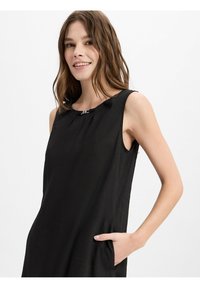 Zwarte mouwloze jurk met een ronde hals, voorzien van een zilveren accent en zijzakken. Gemaakt van lichte stof met een lichte textuur.