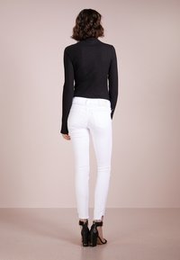 Jersey de cuello alto de color negro con textura acanalada, diseño ajustado; vaqueros skinny blancos; botas de tobillo negras de tacón alto con detalles metálicos. Vista trasera.