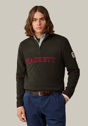 Junger Mann mit schulterlangem braunem Haar, der einen dunkelgrünen Hackett-Pullover, ein helles Hemd darunter und blaue Hosen mit braunem Gürtel trägt.