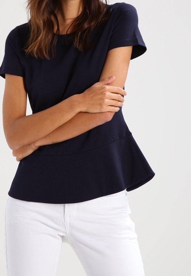 Femme portant un haut ajusté bleu marine à manches courtes avec un basque et un pantalon blanc, bras croisés sur le torse.