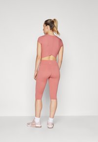 Ensemble de sport rose comprenant un top court avec une découpe dans le dos et un legging capri taille haute. Le matériau semble élastique avec une texture lisse.