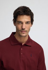 Vinröd pikétröja med krage, tre knappar och en liten blå broderad logotyp på vänster bröst, gjord av texturerad bomull.