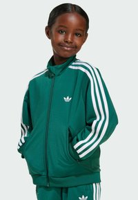 Zelena zip-up jakna od glatkog materijala sa bijelim trakama duž rukava i logom. Dva prednja džepa, visoki ovratnik, opušteni kroj.