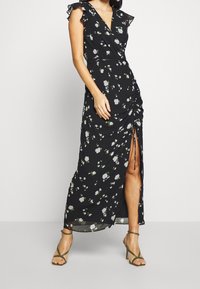 Žena v černých květinových šatech s volány a vysokým rozparkem, která nosí olivově zelené sandály na vysokém podpatku, stojí na bílé podlaze.