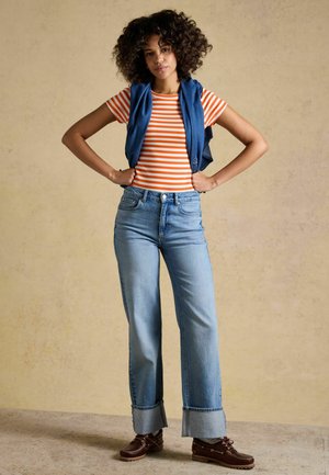 Moteris su garbanotais plaukais stovi rankas ant klubų, dėvi oranžinę ir baltą dryžuotą marškinėlį, mėlynas džinsus, mėlyną megztinį užmėtusi ant pečių, rudos spalvos batus.